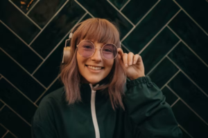 A young woman with red hair, sunglasses, and headphones smiling in front of a green tiled wall.